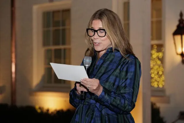 Woman in glasses and plaid shirt reads from paper at microphone