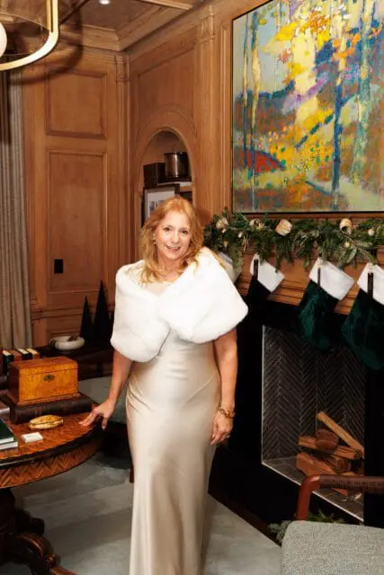 Woman in a champagne dress and white fur stole by a fireplace with Christmas stockings.