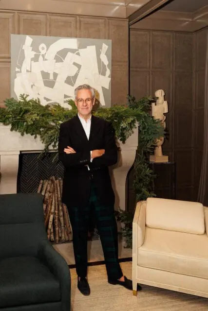 Man in black jacket and plaid pants stands by fireplace with Christmas greenery.