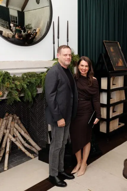 Man in suit and woman in brown dress posing by fireplace with greenery and logs
