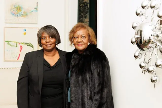 Two women, one in a black blazer and the other in a fur coat, stand indoors.