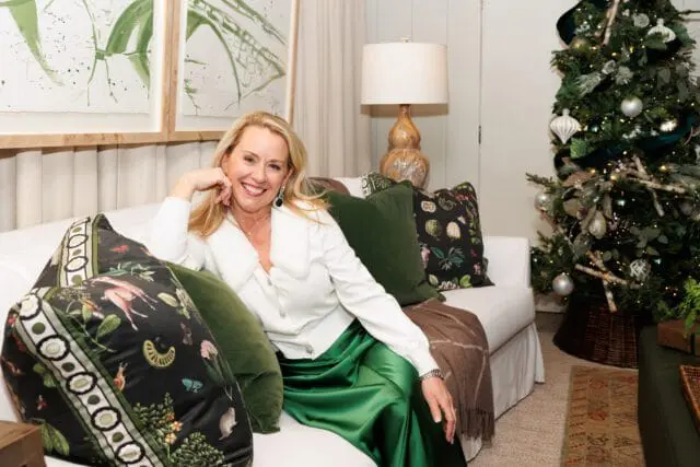 Woman in white cardigan and green skirt sitting on white sofa next to Christmas tree.