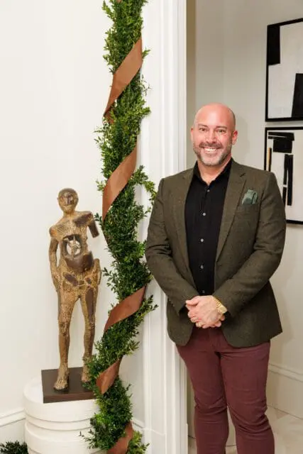 Man in green jacket and maroon pants standing next to greenery and sculpture.