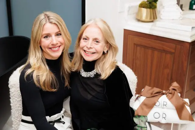 Two women smiling, one wearing a black turtleneck and jeweled necklace, the other a black