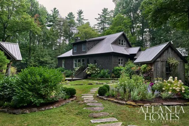 Dark shingled house with metal roof, surrounded by lush gardens and trees.