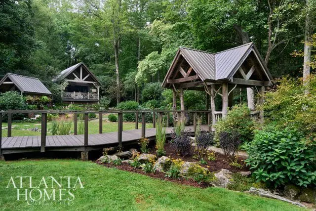 Wooden bridge leads to rustic cabins and gazebo surrounded by lush trees and landscaping.