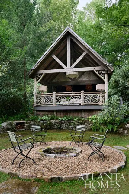 Rustic wooden pavilion with stone fire pit and seating in a garden setting.