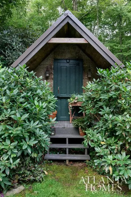 Gable-roofed shed with dark green Dutch door, surrounded by lush green foliage.