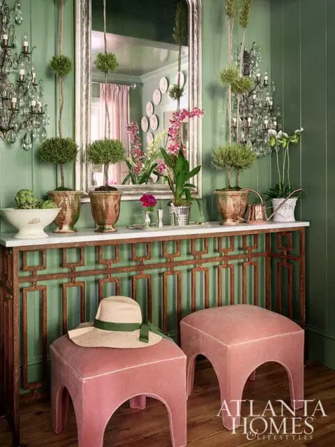 Green paneled dining room with marble console table, pink ottomans, and topiaries.