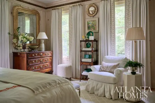 Primary bedroom with antique dresser, white armchair, and Atlanta Homes & Lifestyles logo.