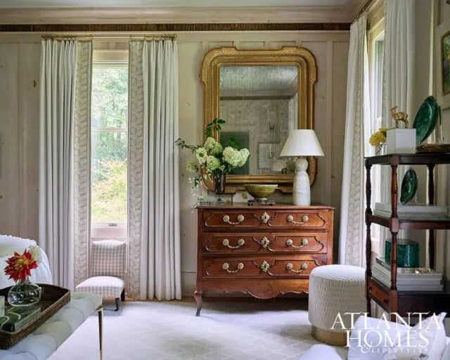 Primary bedroom with wood paneled walls, ornate mirror, antique dresser, and floral curtains.