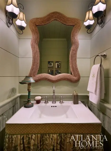 Powder room with green paneled walls, white sink, wicker mirror, and brass sconces.