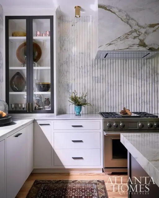 White kitchen with fluted marble backsplash, marble countertops, and glass-front cabinets.