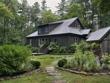Dark wood shingle house with metal roof surrounded by lush gardens and trees. Stone path leads to
