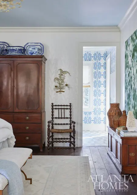 Wooden armoire with blue and white plates, spindle chair, and blue patterned wallpaper.