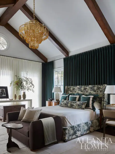 Bedroom with vaulted ceiling, exposed beams, gold chandelier, and dark green curtains.