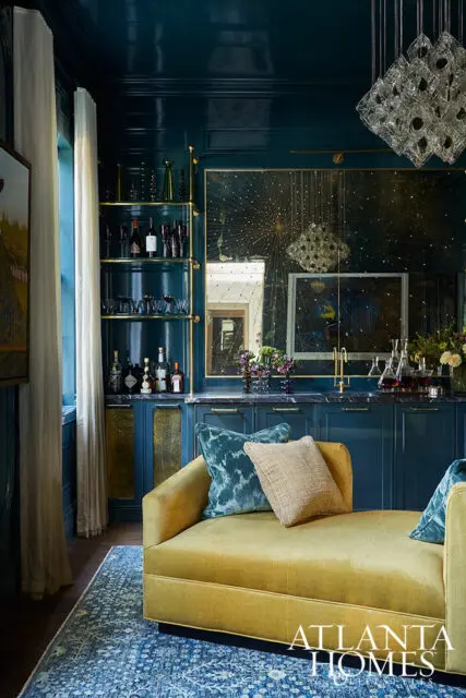 Dark teal bar with mirrored backsplash, gold shelving, and yellow chaise lounge.