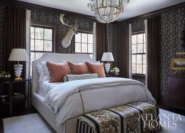Bedroom with dark floral wallpaper, white bed with coral pillows, and taxidermy deer head.