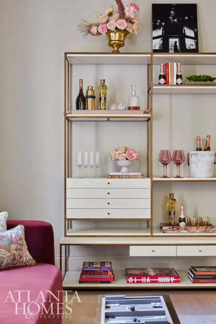 White shelving unit with gold trim holding barware, champagne bottles, books, and floral