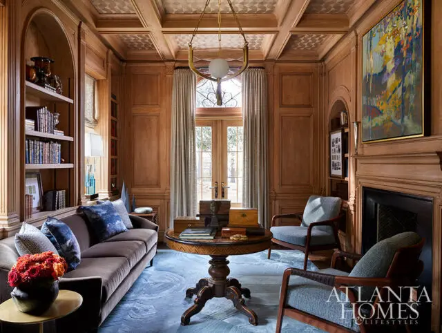 Wood-paneled library with a brown sofa, two armchairs, a round table, and a fireplace.