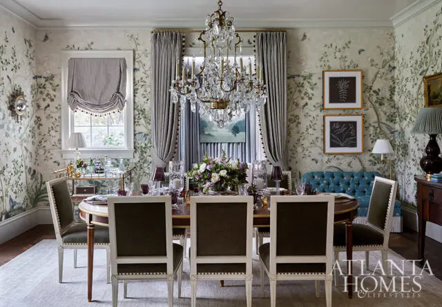 Dining room with floral wallpaper, oval table, dark upholstered chairs, and crystal chandelier.