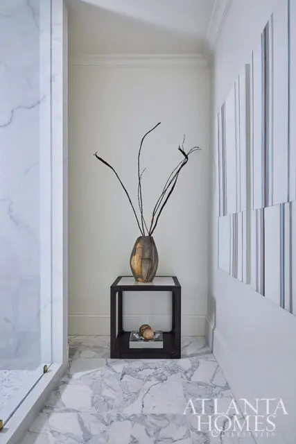 Marble floor and shower surround, modern side table with vase and branches, abstract art on wall.