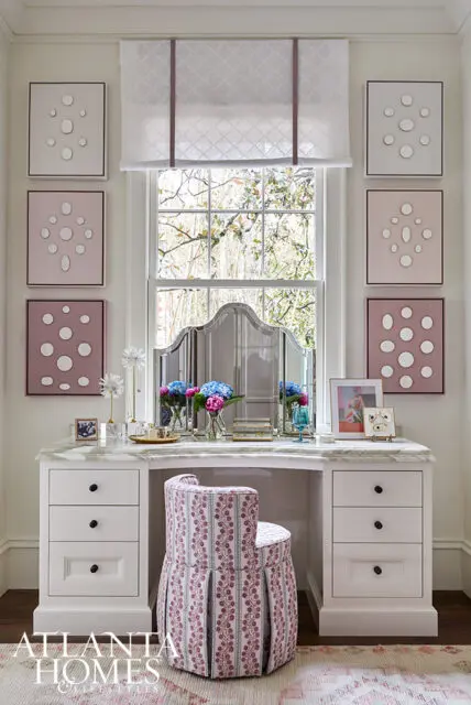 Vanity with marble top, white drawers, and upholstered stool. Framed art on wall.