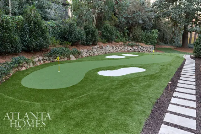Putting green with sand traps and flagsticks, bordered by stone wall and stepping stone path