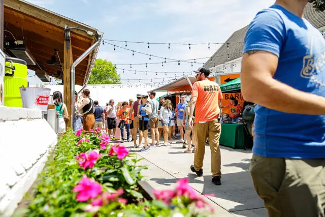 People gathered outdoors at a food event with string lights and flowers in the foreground.