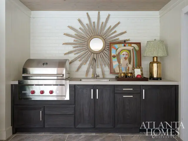 Outdoor kitchen with stainless steel grill, dark wood cabinets, sunburst mirror, and abstract art.