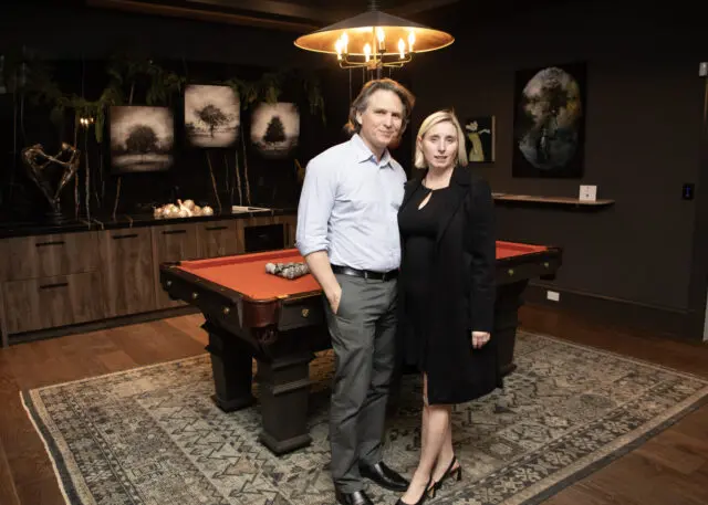 Man and woman stand by a pool table in a room with dark walls and wood cabinetry.