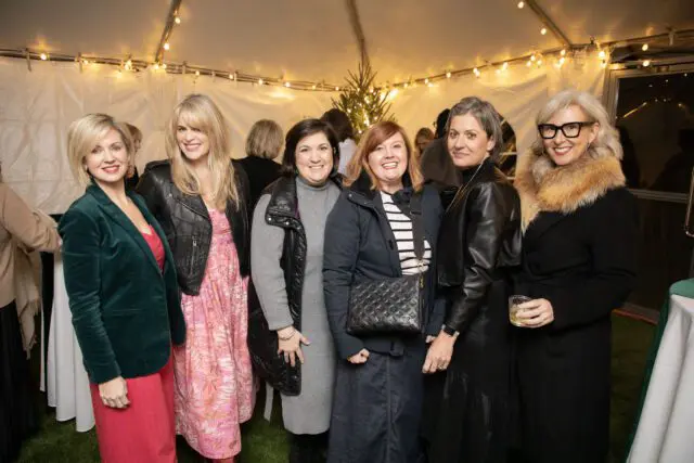 Six women in fashionable attire posing for a photo under a tent with string lights.