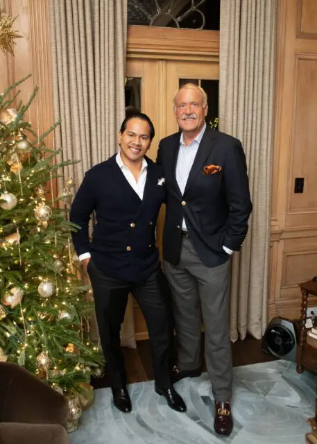 Two men standing next to a decorated Christmas tree in a room with wood paneling and curtains.