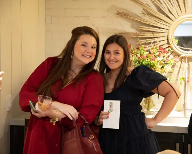 Two women smiling, one in a red dress holding a drink, the other in a navy dress holding a card.