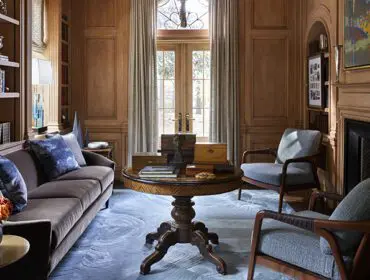 Library with brown sofa, blue accent pillows, ornate round table, and two armchairs.