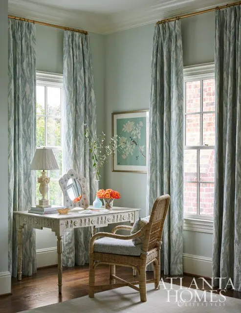 Desk with wicker chair, patterned curtains, and artwork on pale green walls.