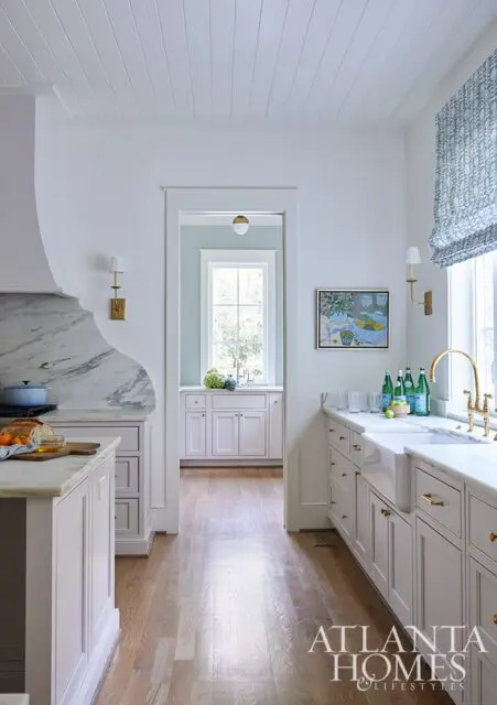 White kitchen with marble countertops, apron sink, brass hardware, and a roman shade.
