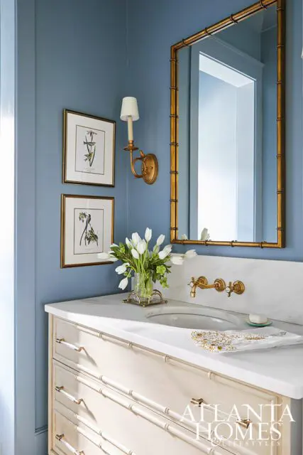 Blue bathroom with marble vanity, bamboo-style drawers, gold mirror, and botanical prints