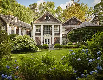 Shingle-style house with white columns and a circular window above the entrance. Lush landscaping