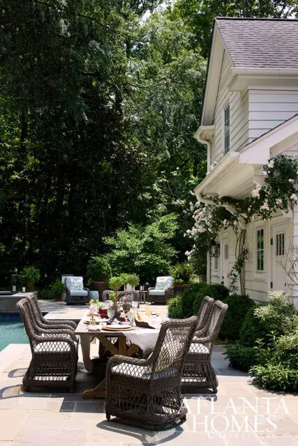 Wicker dining set on stone patio next to pool and white house with climbing roses