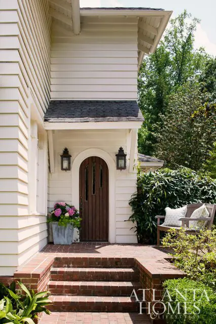 Cream siding house with arched wooden door, brick steps, and porch bench with pillows.