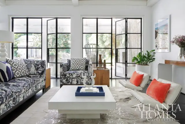 Living room with patterned sofa, white chairs with orange pillows, and large windows.