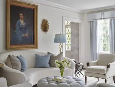 Living room with white sofa, blue pillows, tufted ottoman, and portrait painting.