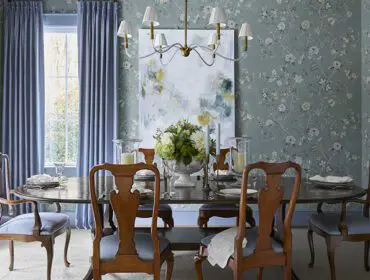 Dining room with floral wallpaper, blue drapes, oval table, and wooden chairs.