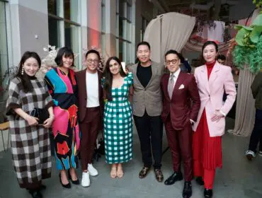Seven people in stylish attire pose for a photo indoors.