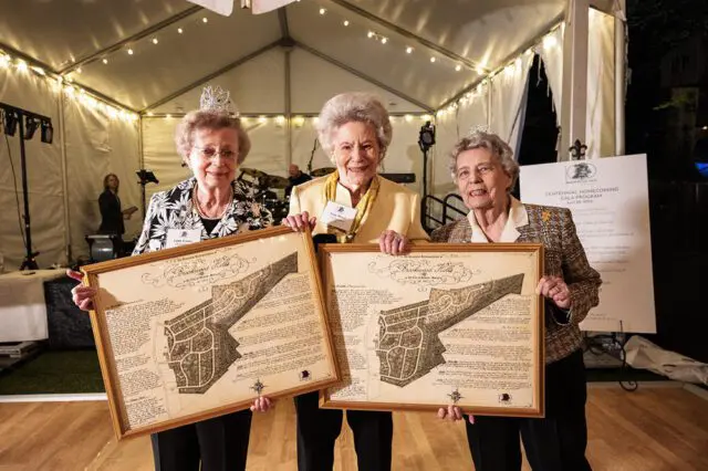 Three women in tiaras holding framed maps of Brookwood Hills.