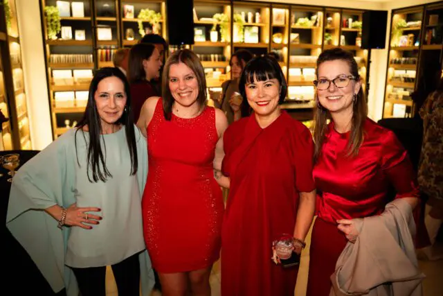 Four women smiling, two in red dresses, one in a light blue top, and one in a red satin top holding