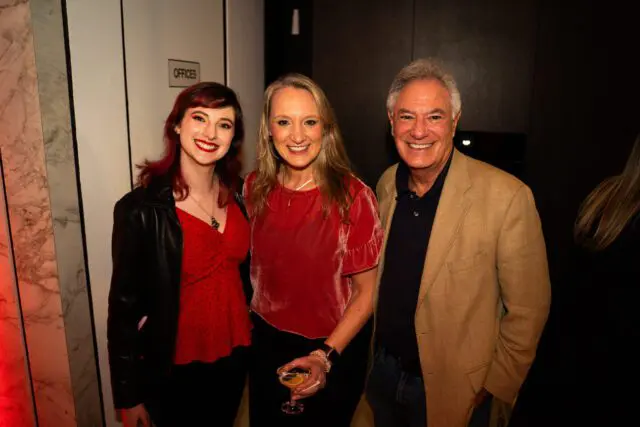 Three people smiling, two women and one man. The woman on the left has red hair and a black leather