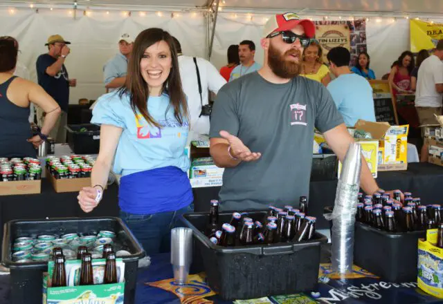 Two people behind a table with coolers of beer bottles and cans.