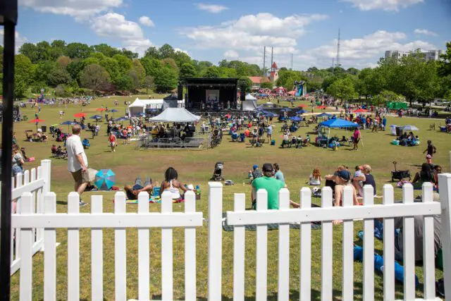 Outdoor festival with a stage, tents, and people relaxing on grass under umbrellas.